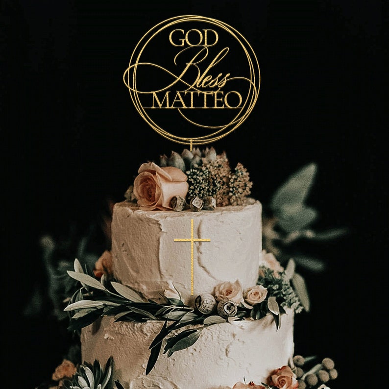 Peut inclure: Un g&acirc;teau blanc &agrave; deux &eacute;tages d&eacute;cor&eacute; de verdure et de fleurs. Un ornement dor&eacute; avec le texte "God Bless Matteo" est plac&eacute; sur le dessus du g&acirc;teau.