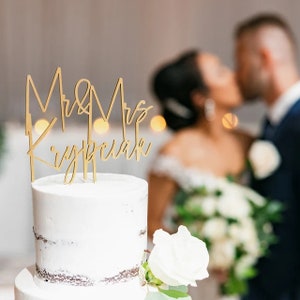 May include: A two-tiered white wedding cake with a gold 'Mr & Mrs Krypciak' topper. The cake is decorated with a brown crumb coating and a white rose.