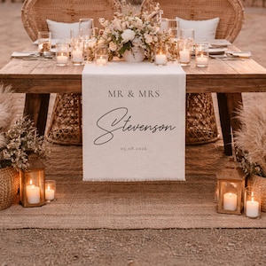 May include: A wedding reception table with a linen table runner displaying "MR & MRS Stevenson 05.08.2026". The table is made of wood and decorated with candles, flowers, and woven chairs, creating a romantic atmosphere.