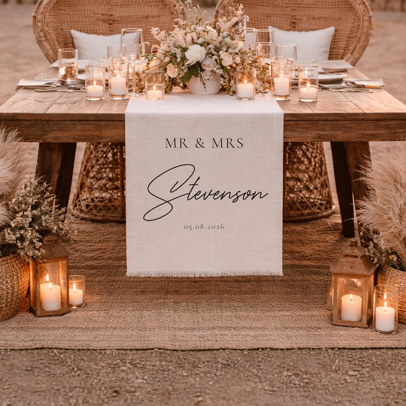 May include: A wedding reception table with a linen table runner displaying "MR & MRS Stevenson 05.08.2026". The table is made of wood and decorated with candles, flowers, and woven chairs, creating a romantic atmosphere.