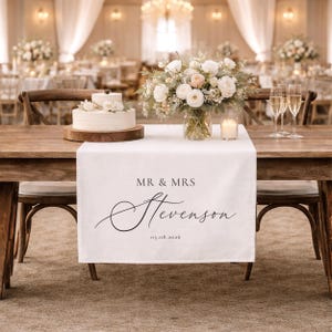 May include: A wedding table setting with a white table runner printed with "MR & MRS Stevenson" and the date 05.08.2026. The table is decorated with a cake, flowers, candles, and champagne glasses. Other tables are visible in the background.