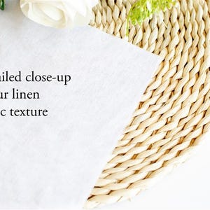 May include: A close-up of white linen fabric with the text "Detailed close-up of our linen fabric texture". The fabric is partially covering a woven, circular placemat. A white flower and greenery are in the background.
