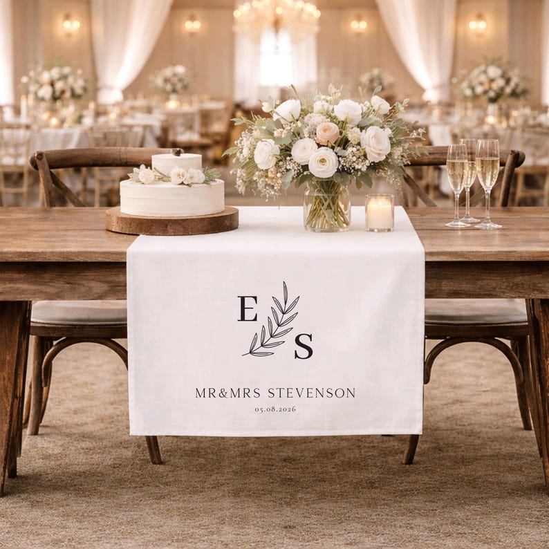May include: A wedding table setting with a white table runner displaying the initials "E" and "S" with a floral motif, and the text "MR&MRS STEVENSON 05.08.2026". The table is adorned with a cake, floral arrangements, and champagne glasses.