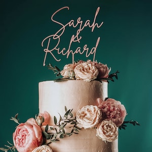 May include: A two-tiered wedding cake decorated with blush pink roses and greenery. The cake topper is a rose gold script that reads "Sarah & Richard".