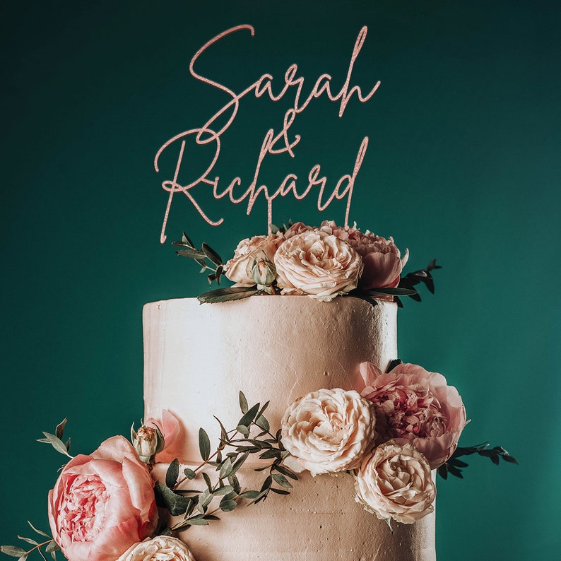 May include: A two-tiered wedding cake decorated with blush pink roses and greenery. The cake topper is a rose gold script that reads "Sarah & Richard".
