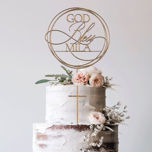 Peut inclure: D&eacute;cor de g&acirc;teau en bois avec un design circulaire et les mots "God Bless Mila". Le d&eacute;cor est sur un g&acirc;teau blanc &agrave; deux &eacute;tages avec des fleurs roses.