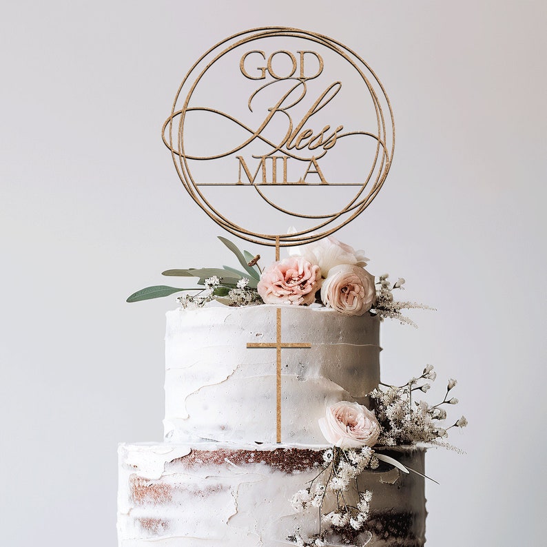Peut inclure: D&eacute;cor de g&acirc;teau en bois avec un design circulaire et les mots "God Bless Mila". Le d&eacute;cor est sur un g&acirc;teau blanc &agrave; deux &eacute;tages avec des fleurs roses.