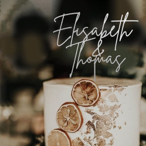 May include: A white cake with dried orange slices and gold leaf accents. The cake is topped with a silver glitter topper that reads "Elisabeth & Thomas".