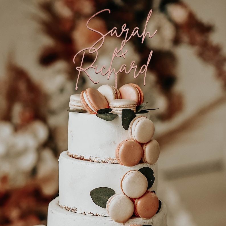 May include: A three-tiered wedding cake decorated with pastel pink and beige macarons and greenery. The cake topper says "Sarah & Richard" in a cursive script.