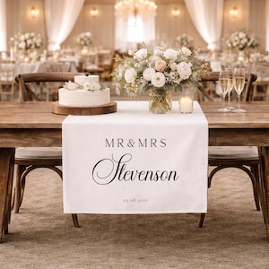 May include: A wedding table setting featuring a white linen runner printed with "MR & MRS Stevenson" and the date 05.08.2026. The table is adorned with a cake, floral arrangements, champagne flutes, and a lit candle. The background shows more tables and floral decorations.