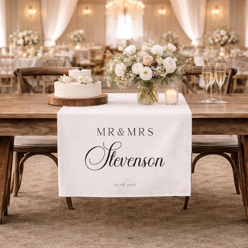 May include: A wedding table setting featuring a white linen runner printed with "MR & MRS Stevenson" and the date 05.08.2026. The table is adorned with a cake, floral arrangements, champagne flutes, and a lit candle. The background shows more tables and floral decorations.