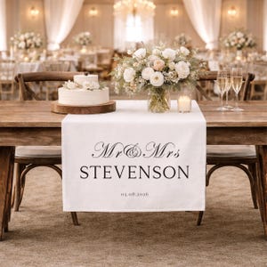 May include: A wedding table setting features a white runner with "Mr & Mrs STEVENSON" and the date printed on it. A two-tiered cake, floral display, and champagne flutes are arranged on the table. The room is decorated with flowers and soft lighting.