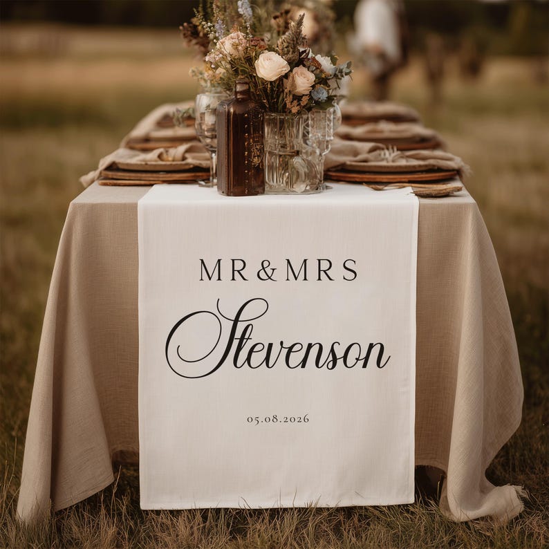 May include: A wedding table setting with a beige tablecloth and a white table runner. The runner has the text "MR & MRS Stevenson" and the date 05.08.2026 in black. The table is set with plates, glasses, and floral arrangements.