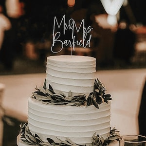 May include: A two-tiered white wedding cake decorated with green leaves and a silver cake topper that reads "Mr & Mrs Barfield".