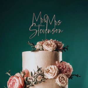 May include: A two-tiered wedding cake decorated with blush pink roses and greenery. The cake topper is a rose gold sign that reads "Mr. & Mrs. Stevenson".