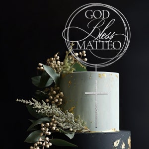 Peut inclure: Un g&acirc;teau &agrave; deux &eacute;tages d&eacute;cor&eacute; d'une croix argent&eacute;e et d'un dessus argent&eacute; qui dit "GOD Bless MATTEO". Le g&acirc;teau est glac&eacute; d'une couleur bleu clair avec des paillettes dor&eacute;es et est entour&eacute; de verdure.