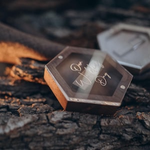 Peut inclure: Une boîte à alliances en bois avec un couvercle transparent et l'inscription "Our Wedding Day". La boîte est posée sur un morceau d'écorce.