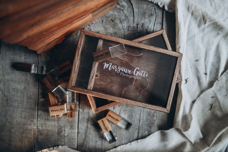 May include: A wooden box with a clear lid, containing a wooden USB drive with the text "Margaux Gatti Photographie" on it. The box is surrounded by other wooden USB drives.