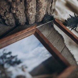 May include: A wooden picture frame with a glass insert, showcasing a photograph of a tree and a cloudy sky. The frame is sitting on a wooden surface with a piece of fabric and dried flowers.