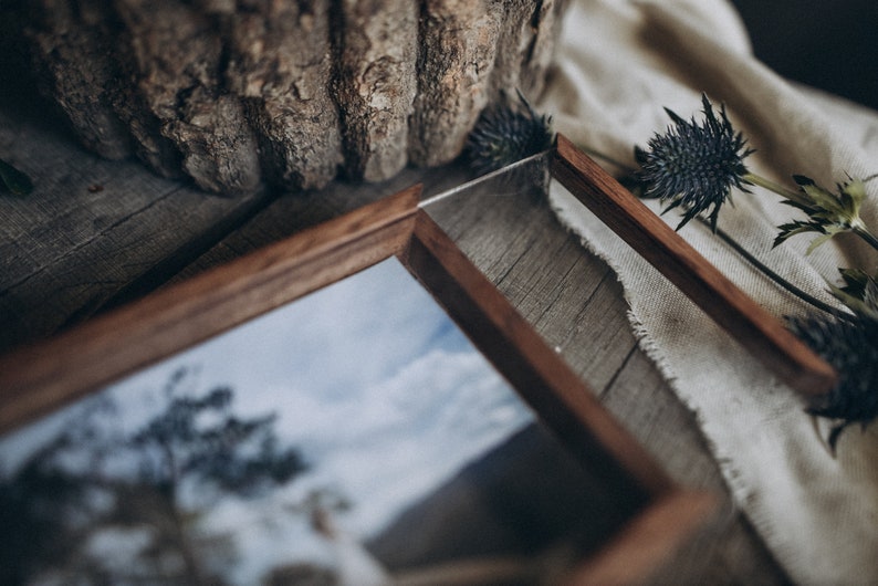 May include: A wooden picture frame with a glass insert, showcasing a photograph of a tree and a cloudy sky. The frame is sitting on a wooden surface with a piece of fabric and dried flowers.
