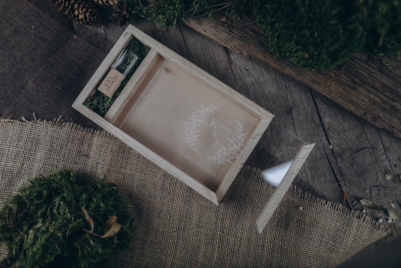 May include: A wooden box with a lid, engraved with a wreath and the initials "K & I" and the date "October 27th, 2023". The box contains a small wooden USB drive with the text "K&I Memories" engraved on it.