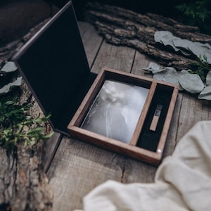 Puede incluir: Una caja de madera con forro de terciopelo negro, que contiene una fotografía y una pequeña unidad USB de madera. La caja está abierta y se encuentra sobre una superficie de madera.
