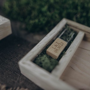 May include: A wooden box with a glass insert containing a small wooden USB drive with the inscription "L&Z 09.30.23" on it. The box is filled with green moss.