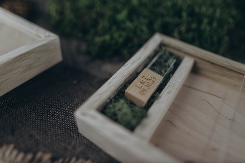 May include: A wooden box with a glass insert containing a small wooden USB drive with the inscription "L&Z 09.30.23" on it. The box is filled with green moss.