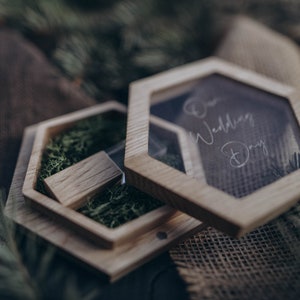 Puede incluir: Una caja hexagonal de madera con una tapa transparente. La tapa tiene las palabras "Our Wedding Day" escritas en ella. La caja está llena de musgo verde y un pequeño bloque de madera.