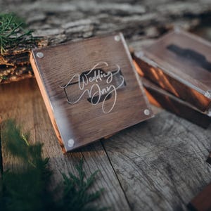 Op de afbeelding: Een vierkante houten doos met een doorzichtig acryldeksel waarop de tekst Wedding Day in witte letters staat. De doos heeft een donkere houtnerf en is voorzien van metalen hoekbevestigingen aan de bovenkant.