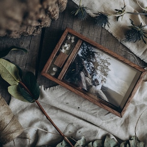 May include: A wooden box with a hinged lid, containing a photograph of a couple standing on a rock overlooking a landscape. The box is decorated with dried flowers and is sitting on a piece of fabric.