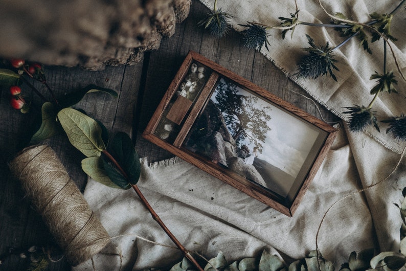 May include: A wooden box with a hinged lid, containing a photograph of a couple standing on a rock overlooking a landscape. The box is decorated with dried flowers and is sitting on a piece of fabric.