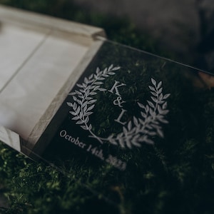 May include: A clear acrylic box with a wooden lid. The box is engraved with the initials "K & L" and the date "October 14th, 2023". The box is surrounded by green moss.