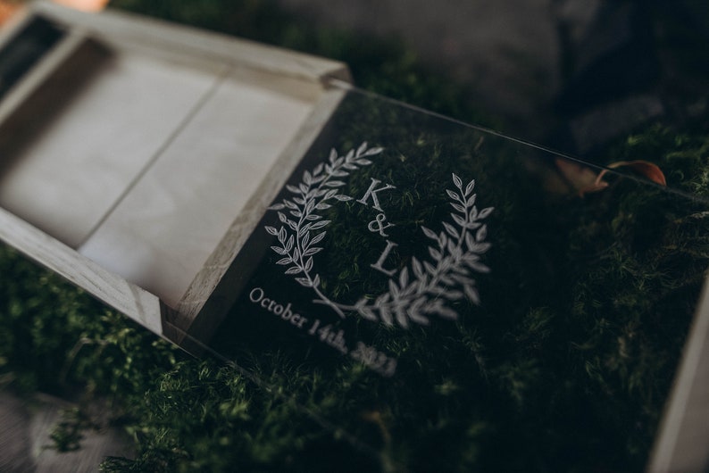 May include: A clear acrylic box with a wooden lid. The box is engraved with the initials "K & L" and the date "October 14th, 2023". The box is surrounded by green moss.