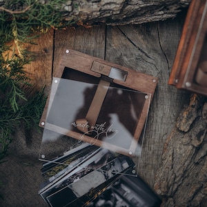 May include: A dark wood photo box with a clear acrylic sliding lid featuring the white script text made with love and a small heart icon. The box contains a stack of rectangular photographs and includes a brown ribbon pull tab for easy access.