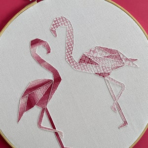 May include: A white embroidery hoop featuring two pink origami flamingo designs. The flamingos are facing each other and are made of geometric shapes.