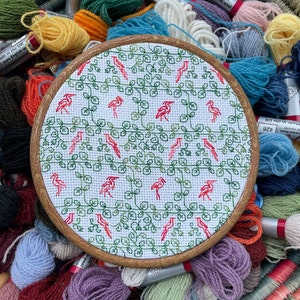 May include: A round embroidery hoop with a white fabric background featuring a repeating pattern of green leaves and red birds. The hoop is surrounded by colourful yarn skeins.