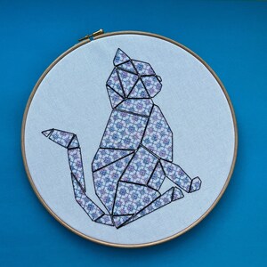 May include: A geometric cat design embroidered on white fabric in a wooden embroidery hoop. The cat is made up of blue and pink patterned shapes.