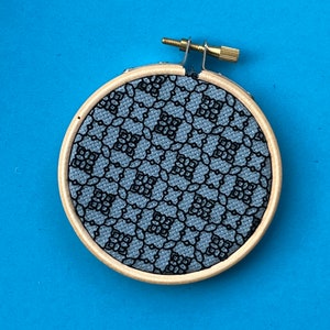 May include: A small, round embroidery hoop with a light wood frame. The fabric inside is a light blue with a black lace pattern. The hoop has a brass screw and hardware for tightening the fabric. The background is a solid blue.