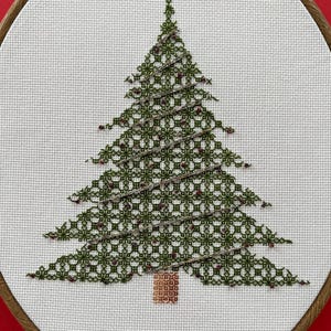 Puede incluir: Bordado de punto de cruz de un árbol de Navidad en un aro de madera. El árbol es verde con un patrón detallado y pequeños acentos rojos. El tronco es marrón. El fondo es blanco y el aro está sobre un fondo rojo.