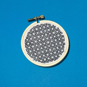 May include: A black and white geometric pattern embroidered on a round white canvas, framed in a natural wood hoop.