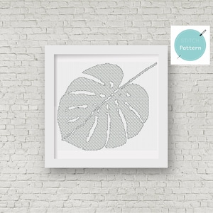 May include: A black and white cross stitch pattern of a monstera leaf in a white frame. The pattern is for a cross stitch project.