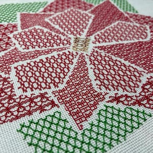 以下が含まれることがあります： ポインセチアの花の赤と緑のクロスステッチ刺繍のクローズアップ。花は赤い糸で詳細なパターンで刺繍されており、葉は緑の糸で刺繍されています。