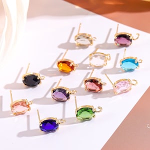 Może przedstawiać: Zestaw 10 owalnych elementów do kolczyków z kamieniami szlachetnymi, ze złotymi sztyftami i pętlami. Kamienie szlachetne są w różnych kolorach, w tym czarny, różowy, zielony, niebieski, fioletowy, pomarańczowy i przezroczysty.