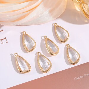 May include: Six clear glass teardrop-shaped pendants with gold-toned metal frames. The pendants are arranged on a white surface.