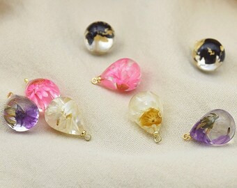 May include: A collection of clear resin teardrop-shaped pendants and spherical beads, each containing dried flowers or gold leaf. The pendants feature various colors of flowers, including pink, purple, and yellow, with gold-colored loops for hanging.