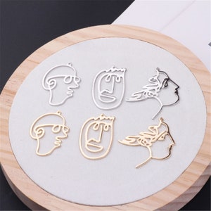 May include: Six silver and gold metal charms in the shape of abstract faces. The charms have a hole at the top for attaching to jewelry.