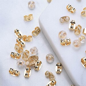 May include: A collection of gold-colored earring backs and clear, rounded earring stoppers are scattered on a white surface. The earring backs have various designs, including butterfly and flat styles. The clear stoppers are small and round.
