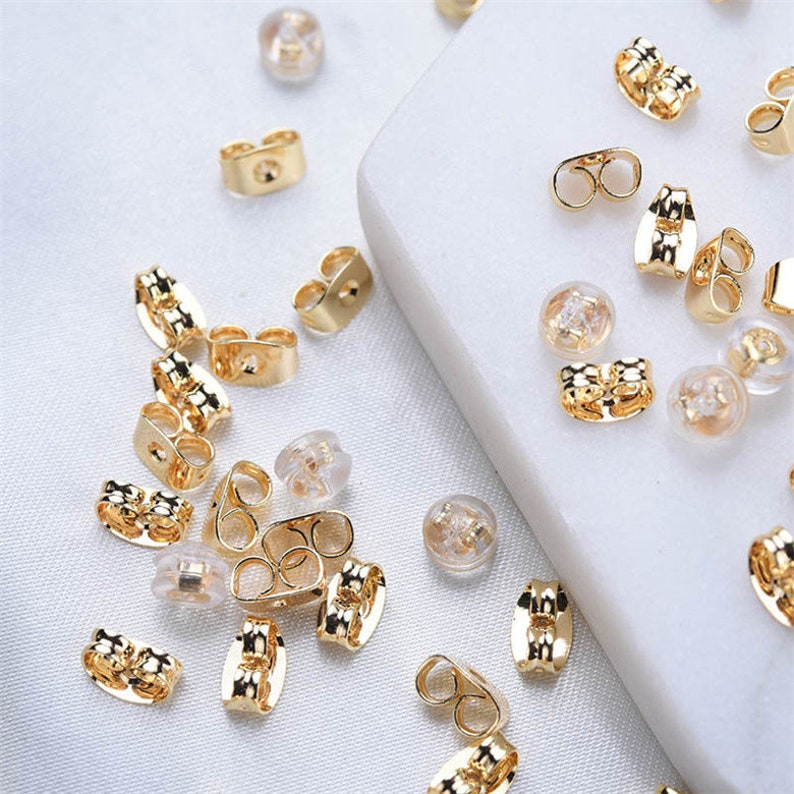 May include: A collection of gold-colored earring backs and clear, rounded earring stoppers are scattered on a white surface. The earring backs have various designs, including butterfly and flat styles. The clear stoppers are small and round.