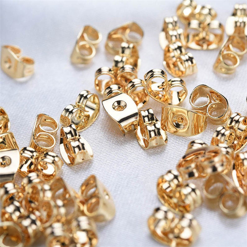 May include: A collection of shiny gold-colored earring backs scattered on a white, textured surface. The earring backs are in various shapes, including butterfly and flat disc styles. The lighting highlights the metallic sheen of the jewelry findings.